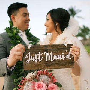 May include: A newly married couple holds a wooden sign that says "Just Maui'd" on a beach. The bride is wearing a white wedding dress and a veil. The groom is wearing a gray suit and a lei. They are standing in front of the ocean.
