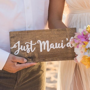 May include: A couple holds a wooden sign that says "Just Maui'd" on a beach. The bride is wearing a white dress and holding a bouquet of colorful flowers.
