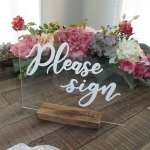 May include: A clear acrylic sign with the text "Please sign" in white cursive lettering. The sign is standing on a wooden base and is surrounded by pink, white, and purple flowers.