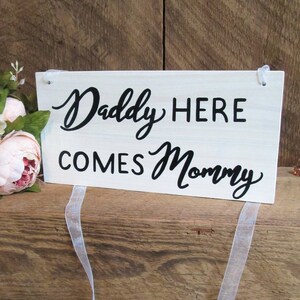 May include: White wooden sign with black text that reads "Daddy Here Comes Mommy" with a white ribbon attached.