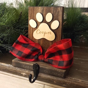 May include: A wooden pet stocking holder with a paw print design and the name "Cooper" engraved on it. It features a red and black buffalo plaid bow and a black hook for hanging. The holder is set against a backdrop of greenery.