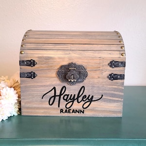 May include: A wooden chest with a curved top and metal hardware. The chest is stained in a light gray color and features the name "Hayley Raeann" in black script on the front. Decorative flowers are in the lower left corner.