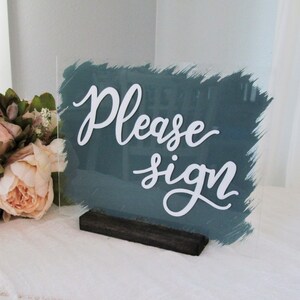 May include: A teal acrylic sign with a white brushstroke background and the text "Please sign" in white cursive lettering. The sign is on a black wooden stand.
