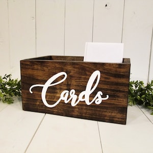 May include: Dark brown wooden card box with white cursive lettering that says 'Cards'.  The box is rectangular and has a rustic look. A white card is visible inside the box. The box sits on a white surface with greenery on either side.