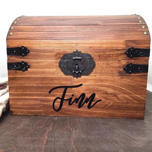 May include: A wooden chest with a curved top, featuring decorative metal hardware and the name "Finn" in black script. The chest is stained a warm brown color and has a vintage aesthetic.