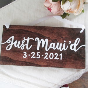 May include: A dark brown wooden sign with white text that reads "Just Maui'd 3-25-2021". The sign is hanging from a white ribbon.