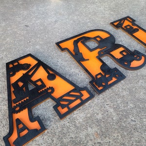 May include: Orange wooden letters spelling out the name "ARIO" with black construction vehicle cutouts.