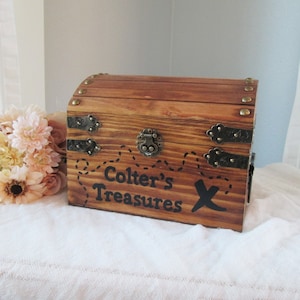May include: A wooden treasure chest with metal hardware and the text "Colter's Treasures" and an "X" on the front. The chest is brown with a curved top and is decorated with brass studs. A bouquet of flowers sits to the left.