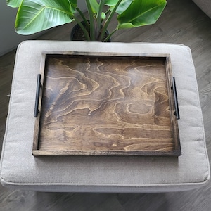 Puede incluir: Una bandeja de servir de madera con asas de metal negro. La bandeja tiene un tinte marr&oacute;n oscuro y un patr&oacute;n de veta de madera. La bandeja est&aacute; sentada sobre una superficie de tela gris claro.