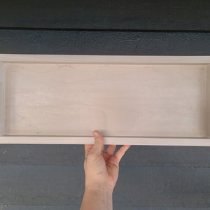 Puede incluir: Una bandeja rectangular de madera blanca con asas de metal negro. La bandeja es lisa.