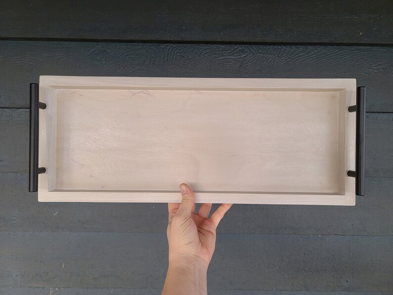 Puede incluir: Una bandeja rectangular de madera blanca con asas de metal negro. La bandeja es lisa.