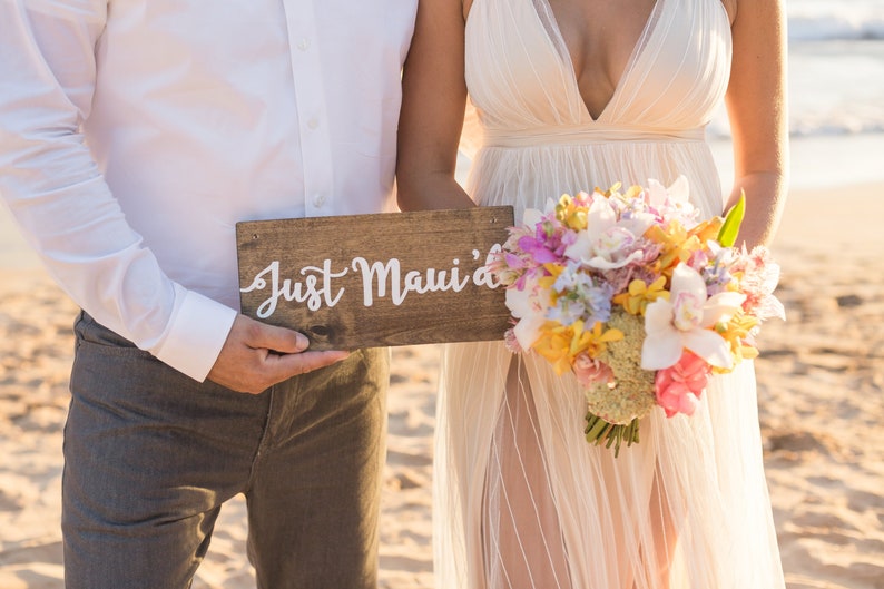 May include: A couple holds a wooden sign that says "Just Maui'd" on a beach. The bride is wearing a white dress and holding a bouquet of colorful flowers.