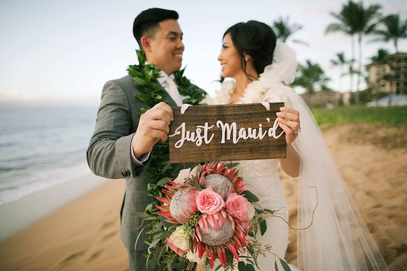May include: A newly married couple holds a wooden sign that says "Just Maui'd" on a beach. The bride is wearing a white wedding dress and a veil. The groom is wearing a gray suit and a lei. They are standing in front of the ocean.