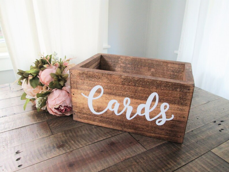 May include: A rustic wooden box with the word "Cards" written in white on the front. The box is sitting on a wooden table with a bouquet of pink flowers next to it.