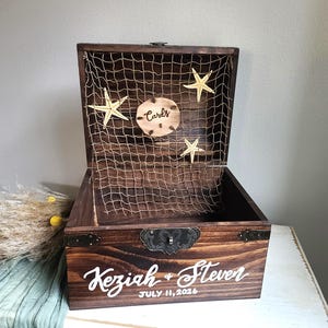 May include: A dark brown wooden box with a hinged lid featuring a fishing net, three starfish, and a circular wooden sign that reads Cards. The front of the box is personalized with the white text Keziah + Steven and the date JULY 11, 2026.