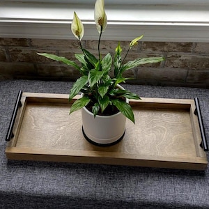 May include: A wooden serving tray with black handles holds a potted peace lily plant. The plant has dark green leaves and white flowers. The tray is light brown and sits on a gray surface near a brick wall.