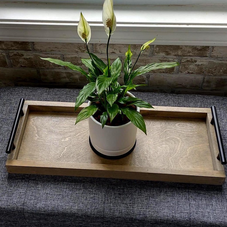 May include: A wooden serving tray with black handles holds a potted peace lily plant. The plant has dark green leaves and white flowers. The tray is light brown and sits on a gray surface near a brick wall.