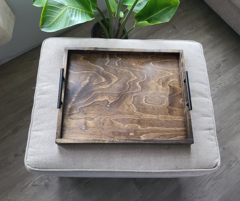Puede incluir: Una bandeja de servir de madera con asas de metal negro. La bandeja tiene un tinte marr&oacute;n oscuro y un patr&oacute;n de veta de madera. La bandeja est&aacute; sentada sobre una superficie de tela gris claro.