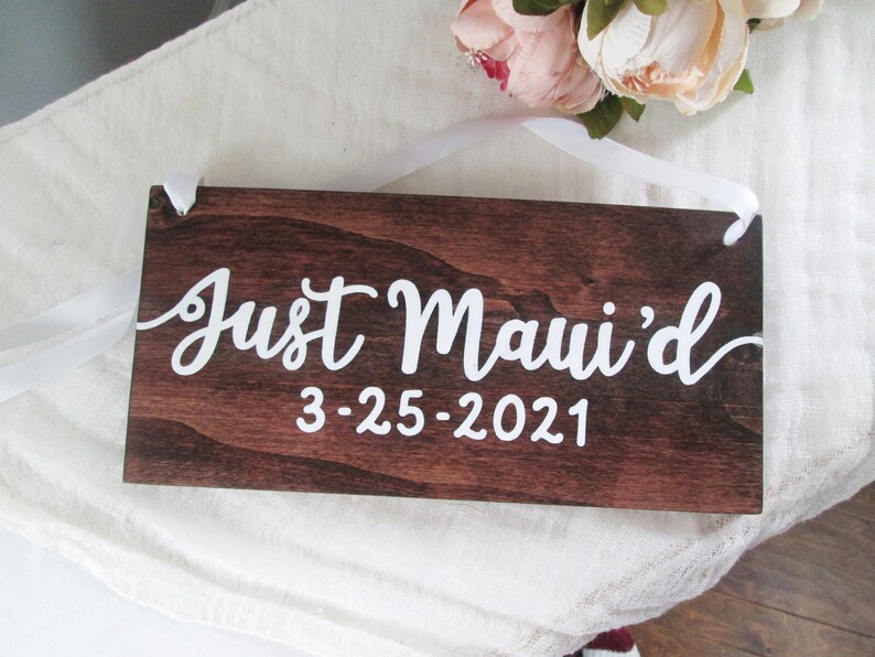 May include: A dark brown wooden sign with white text that reads "Just Maui'd 3-25-2021". The sign is hanging from a white ribbon.