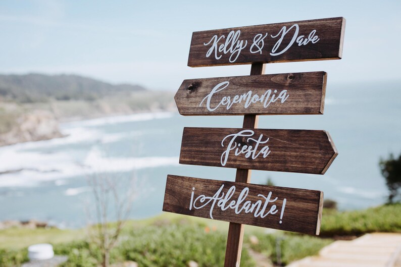 May include: Wooden signpost with four arrows pointing to the right. The top arrow says "Kelly & Dave". The second arrow says "Ceremonia". The third arrow says "Fiesta". The bottom arrow says "&iexcl;Adelante!" The signpost is in front of a blurred background of a beach and ocean.