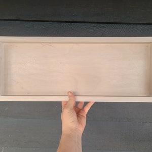 May include: A rectangular wooden serving tray with a light wood finish. It features black metal handles on each end. The tray is held by a hand, showcasing its size and design. Ideal for serving food or displaying items.