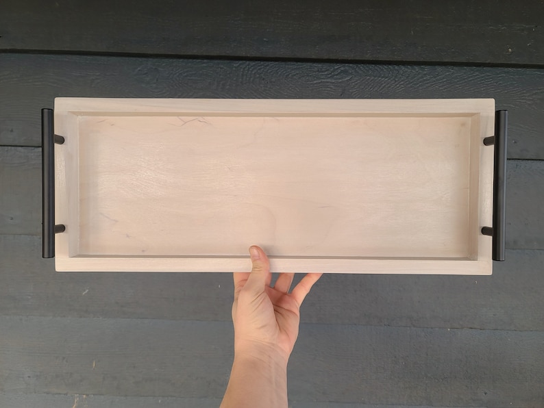 May include: A rectangular wooden serving tray with a light wood finish. It features black metal handles on each end. The tray is held by a hand, showcasing its size and design. Ideal for serving food or displaying items.