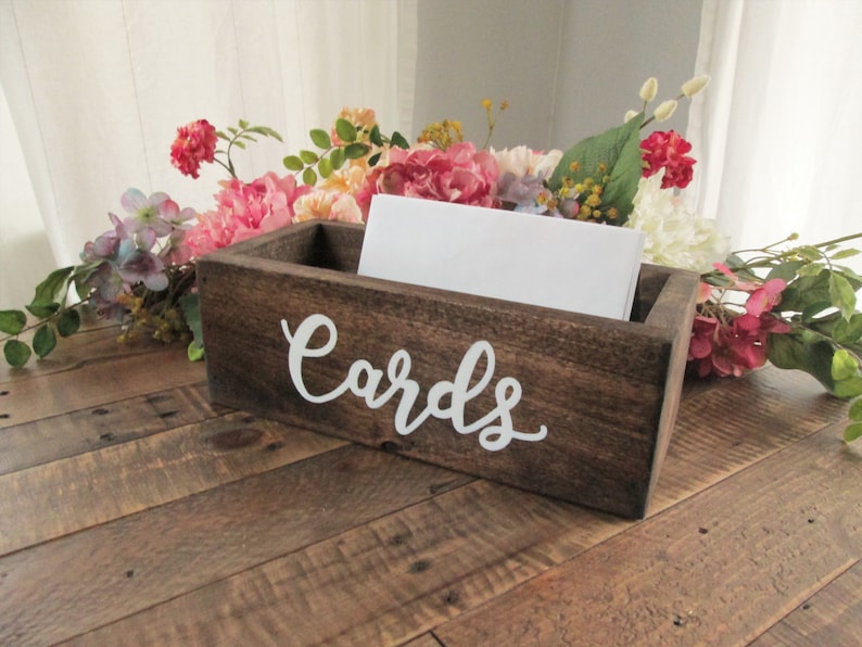 May include: A rustic wooden box with a white inscription that reads "Cards". The box is filled with white envelopes and is surrounded by pink and white flowers.