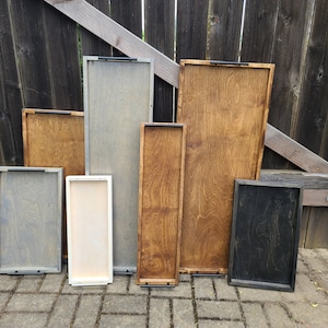 May include: A collection of wooden serving trays in various sizes and finishes. The trays range in color from light beige to dark charcoal, with visible wood grain. Each tray has a dark metal handle.