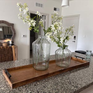 May include: A wooden serving tray with black handles holds two clear glass vases with white flowers. The vases are different sizes and have vertical ribbed detailing. The tray sits on a speckled countertop.