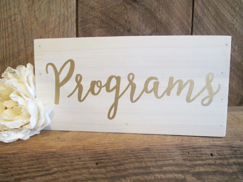 May include: A white wooden sign with the word "Programs" written in gold cursive lettering.