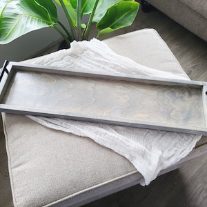 Puede incluir: Una bandeja de madera gris con asas de metal negro. La bandeja est&aacute; sobre una superficie de tela gris claro.