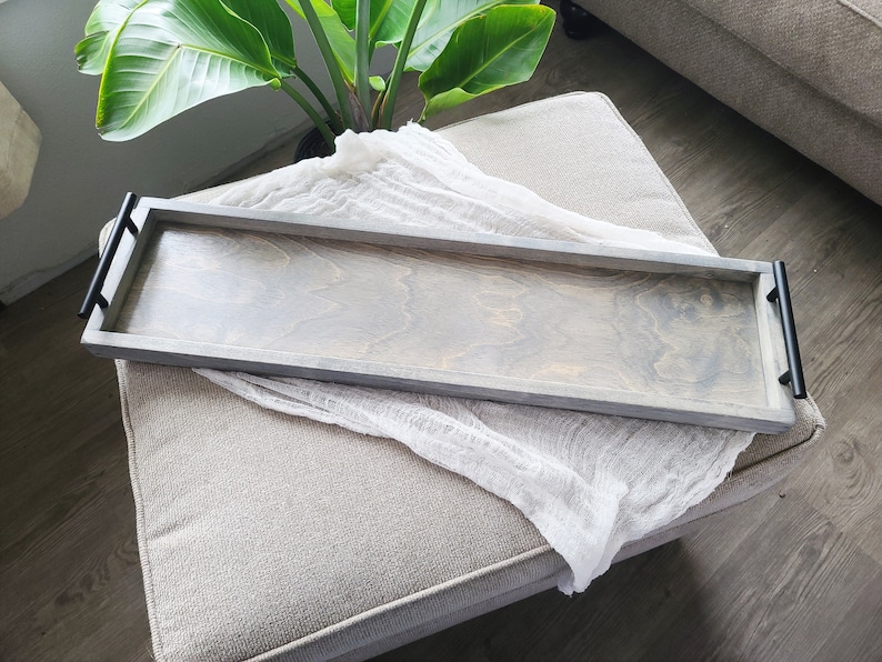 Puede incluir: Una bandeja de madera gris con asas de metal negro. La bandeja est&aacute; sobre una superficie de tela gris claro.