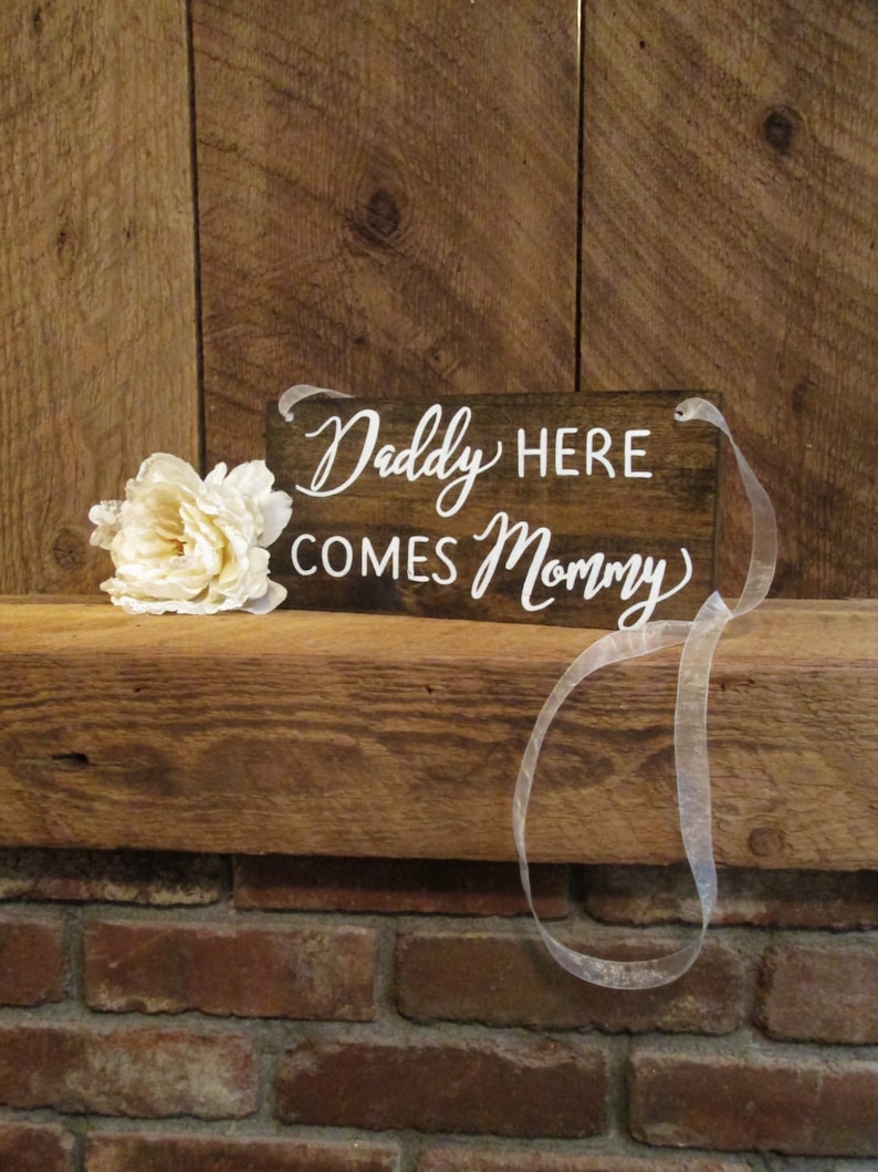 Op de afbeelding: Een rustiek houten bord met witte letters waarop staat "Daddy Here Comes Mommy". Het bord is vastgebonden met een wit lint en ligt op een houten oppervlak.
