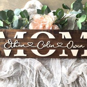 May include: A wooden sign with the word "MOM" in large, light-colored letters. Below, the names "Ethan", "Colin", and "Owen" are displayed in a cursive font, connected by hearts. The sign is set against a backdrop of greenery and fabric.