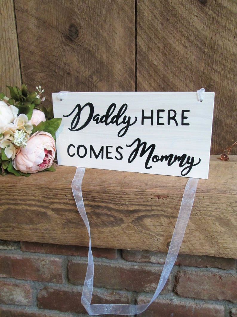 May include: White wooden sign with black text that reads "Daddy Here Comes Mommy" with a white ribbon attached.