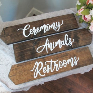 May include: Three wooden arrow signs with white calligraphy. The top sign reads "Ceremony", the middle sign reads "Animals", and the bottom sign reads "Restrooms".