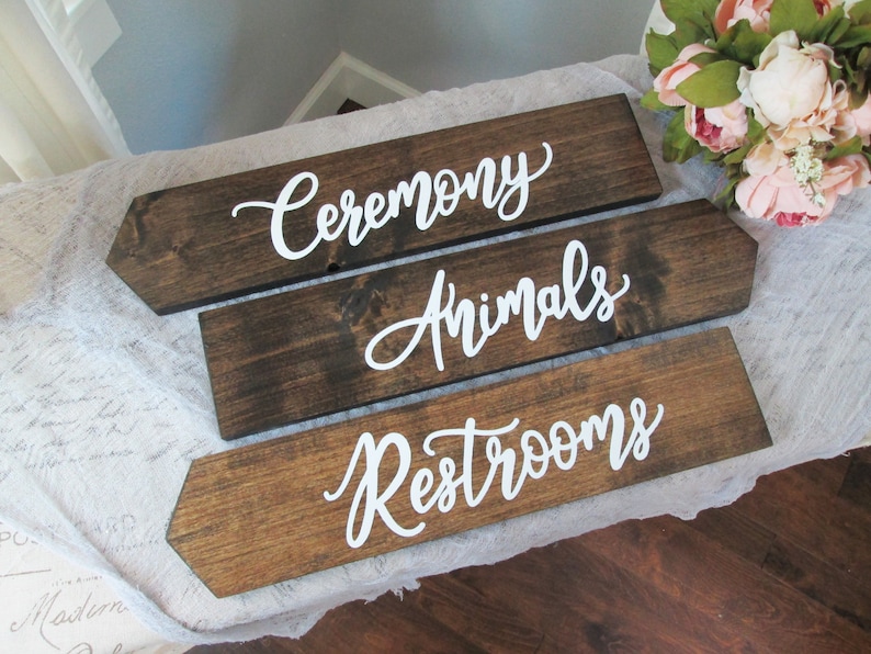 May include: Three wooden arrow signs with white calligraphy. The top sign reads "Ceremony", the middle sign reads "Animals", and the bottom sign reads "Restrooms".