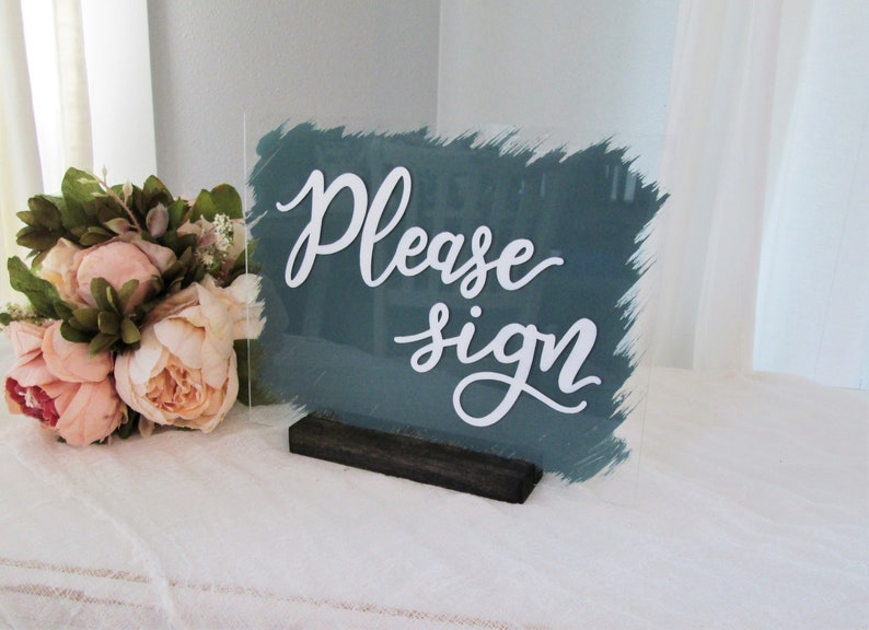 May include: A teal acrylic sign with a white brushstroke background and the text "Please sign" in white cursive lettering. The sign is on a black wooden stand.