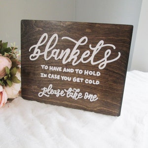 May include: A wooden sign with white cursive text that reads "blankets to have and to hold in case you get cold please take one." The sign is dark brown and sits on a white surface with a bouquet of pink flowers in the background.