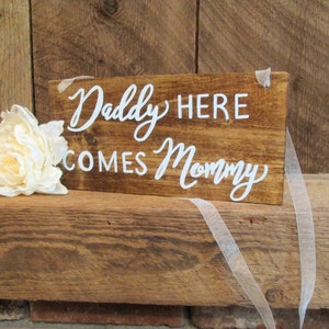 Op de afbeelding: Een rustiek houten bord met witte letters waarop staat "Daddy Here Comes Mommy". Het bord is versierd met een witte bloem en een wit lint.