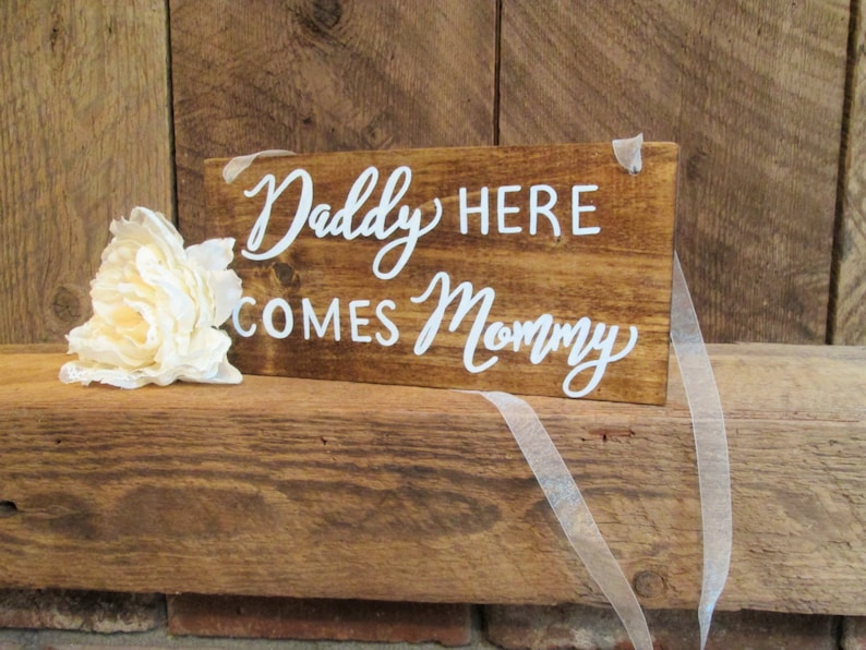 Op de afbeelding: Een rustiek houten bord met witte letters waarop staat "Daddy Here Comes Mommy". Het bord is versierd met een witte bloem en een wit lint.