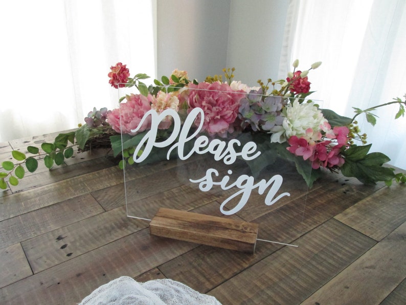 May include: A clear acrylic sign with the text "Please sign" in white cursive lettering. The sign is standing on a wooden base and is surrounded by pink, white, and purple flowers.