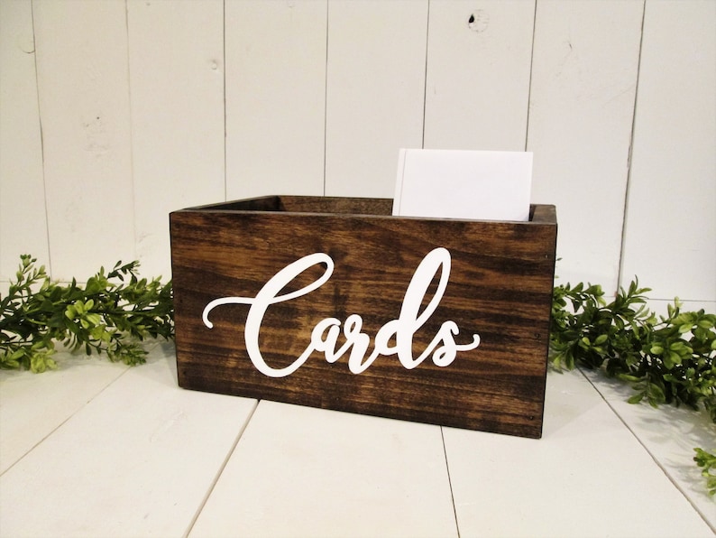 May include: Dark brown wooden card box with white cursive lettering that says 'Cards'.  The box is rectangular and has a rustic look. A white card is visible inside the box. The box sits on a white surface with greenery on either side.