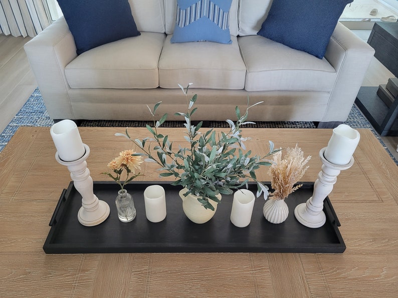 Puede incluir: Una bandeja negra con velas blancas, un jarr&oacute;n con una planta verde, una peque&ntilde;a botella de vidrio y un peque&ntilde;o jarr&oacute;n de cer&aacute;mica blanca con flores secas sobre una mesa de madera.