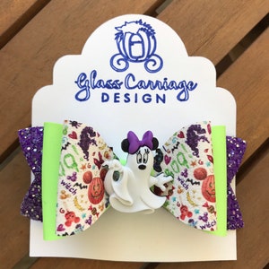 May include: A purple and green glitter hair bow with a white ghost and a Minnie Mouse character with a purple bow. The bow has a white background with a Halloween print of pumpkins, candy corn, and the word "witch".