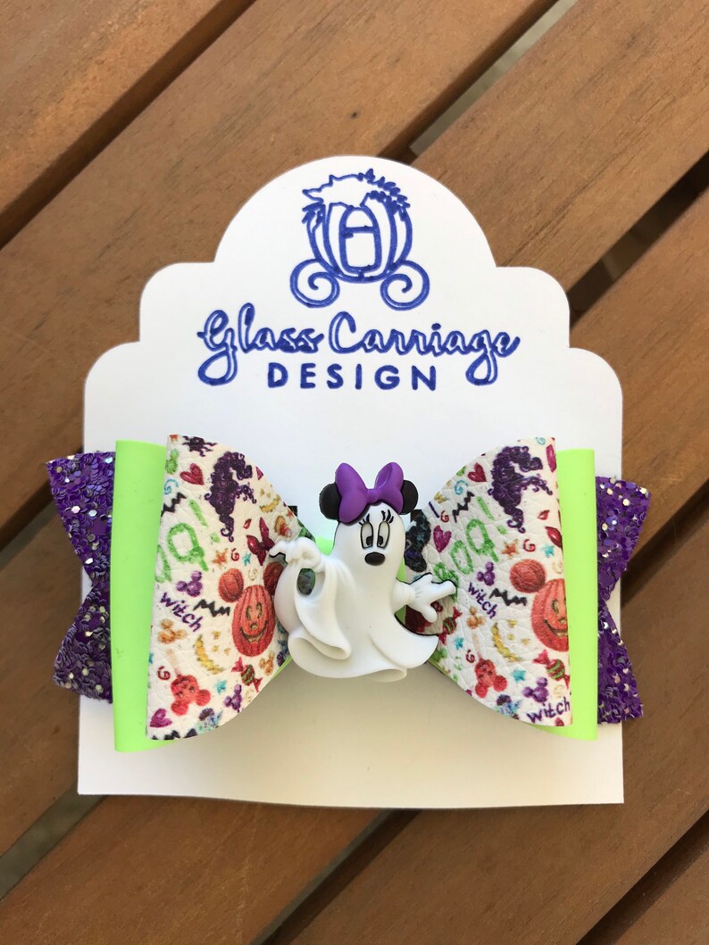 May include: A purple and green glitter hair bow with a white ghost and a Minnie Mouse character with a purple bow. The bow has a white background with a Halloween print of pumpkins, candy corn, and the word "witch".