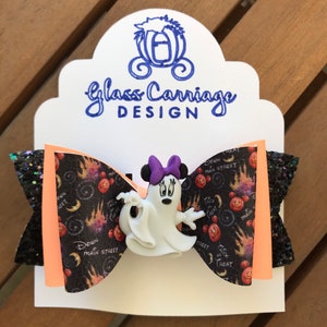 May include: A black and orange Halloween-themed hair bow with a white ghost Minnie Mouse character in the center. The bow has a black glitter fabric on one side and a black and orange fabric with a "Trick or Treat" and "Down Main Street" pattern on the other side.