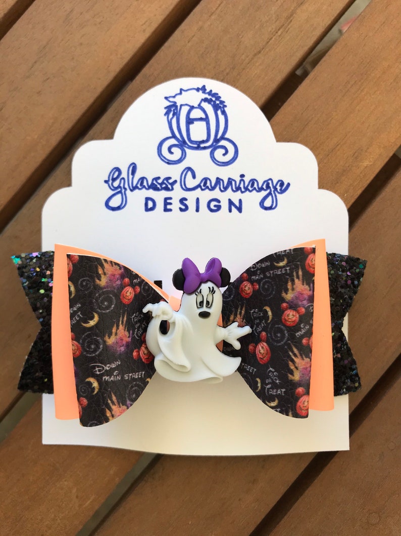 May include: A black and orange Halloween-themed hair bow with a white ghost Minnie Mouse character in the center. The bow has a black glitter fabric on one side and a black and orange fabric with a "Trick or Treat" and "Down Main Street" pattern on the other side.