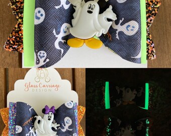 Puede incluir: Un lazo negro y blanco con un fantasma que brilla en la oscuridad y un personaje de Mickey Mouse. El lazo tiene un borde de purpurina verde y naranja.  "Glass Carriage Design" está impreso en una tarjeta blanca detrás del lazo.