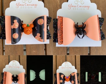 Puede incluir: Dos lazos para el pelo con temática de Halloween con características que brillan en la oscuridad. Un lazo presenta una araña negra y naranja, el otro presenta una calabaza de Mickey Mouse. Ambos lazos están en una tarjeta blanca con el texto "Glass Carriage Design".